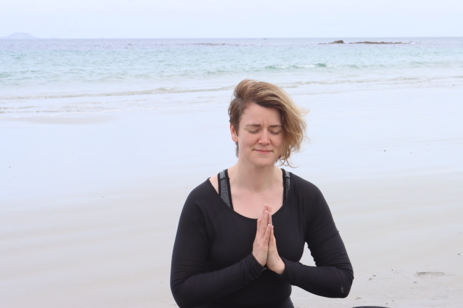 Nicole sitzt am Meer mit geschlossenen Augen, die Hände vor dem Herzen gefaltet