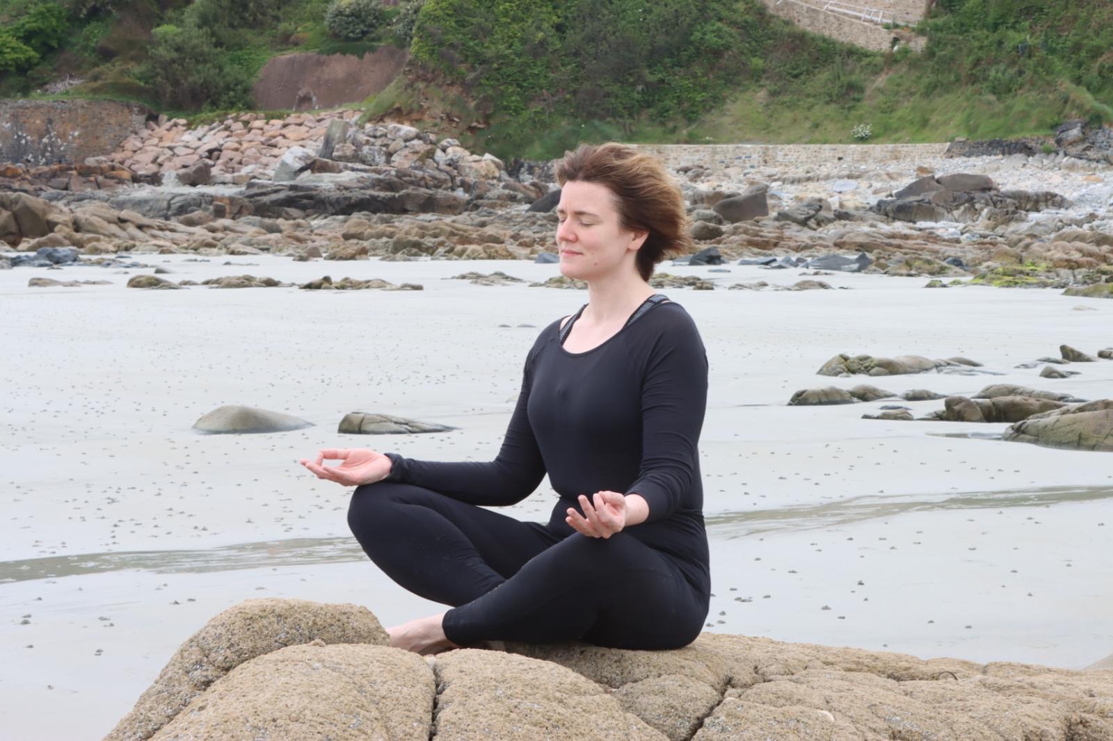 Nicole sitzt am Meer auf einem Felsen, die Augen geschlossen, die Hände entspannt geöffnet auf den Knien